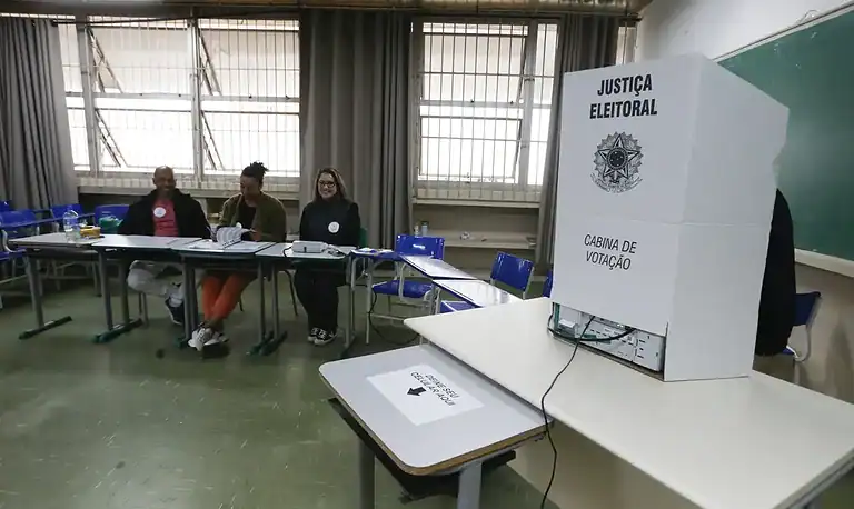 Seção eleitoral com urnas e mesários em sala de votação.